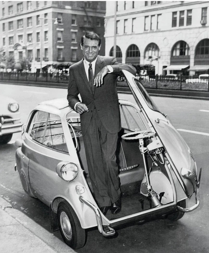The image shows a black and white photograph of the BMW Isetta, known as the smallest BMW ever produced. The Isetta is a tiny, egg-shaped microcar with a front-opening door.