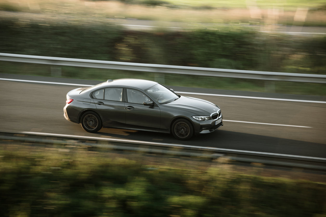 To zdjęcie przedstawia sedana BMW serii 3 poruszającego się po autostradzie. Samochód jest ciemnego koloru, prawdopodobnie szarego lub czarnego i jest lekko rozmazany w wyniku ruchu. To dynamiczne ujęcie podkreśla możliwości samochodu.