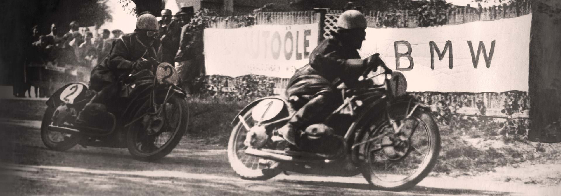 Vintage černobílá fotografie raných motocyklových závodů BMW. Dva jezdci na motocyklech jsou zobrazeni, jak závodí na trati, s transparentem BMW v pozadí.