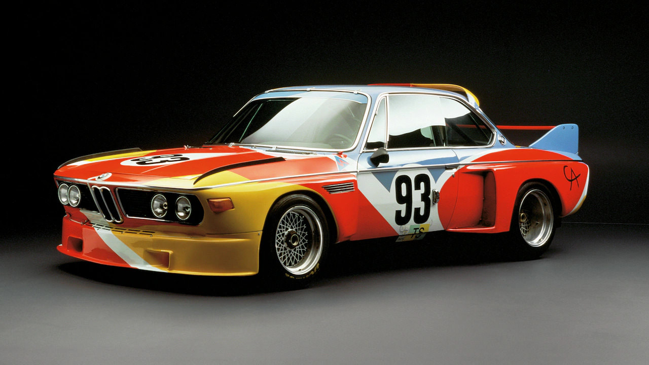 Kolorowy samochód wyścigowy BMW 3.0 CSL z 1975 roku, znany jako Art Car. Pojazd ma efektowny lakier przedstawiający odważne geometryczne kształty w kolorze czerwonym, pomarańczowym, żółtym i niebieskim na białej podstawie.