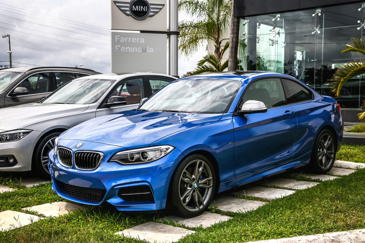 Na pierwszym planie wyraźnie widać jasnoniebieskie coupe BMW serii 2. Samochód stoi na trawie przed salonem samochodowym.