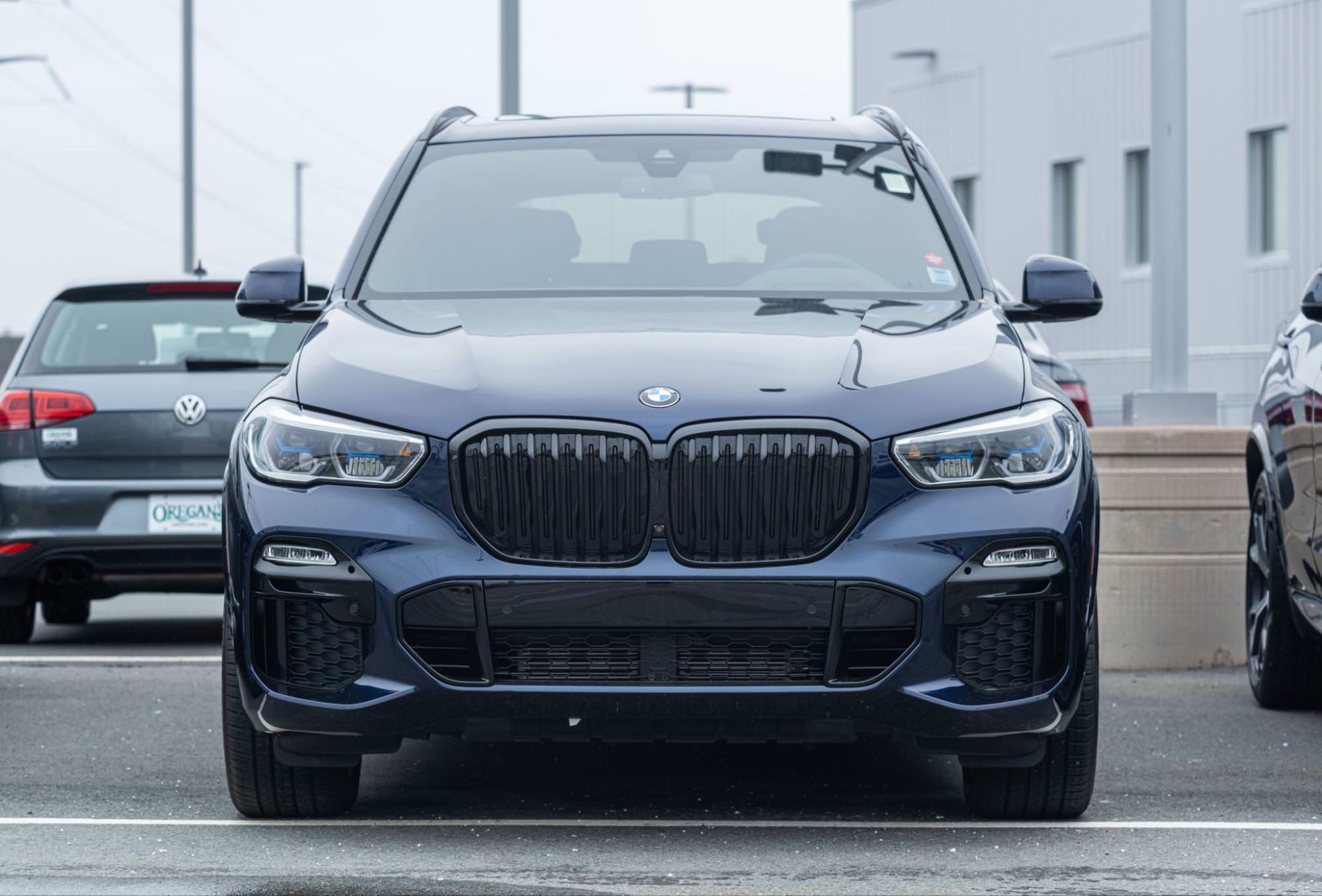 To zdjęcie przedstawia widok z przodu SUV-a BMW X5. Pojazd jest koloru ciemnoniebieskiego lub czarnego i stoi na czymś, co przypomina parking. Zdjęcie koncentruje się na przodzie samochodu, podkreślając jego dużą atrapę chłodnicy.