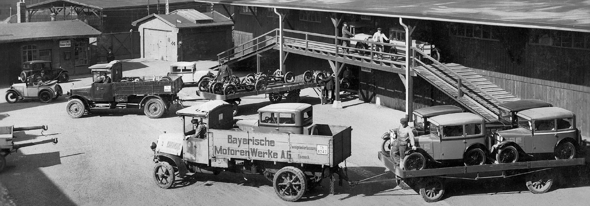 Vintage photograph of the Fahrzeugfabrik Eisenach factory in Germany, showing various early BMW Dixi cars and trucks on display or being loaded onto a ramp.