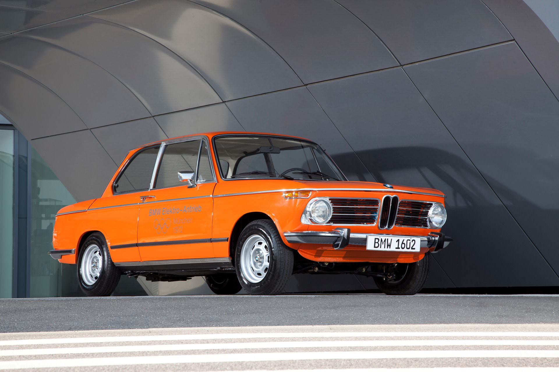 An orange BMW 1602 electric vehicle from 1972 is displayed in a modern setting with curved white walls. The car has a classic, boxy design typical of 1970s sedans, with round headlights and a distinctive BMW kidney grille.