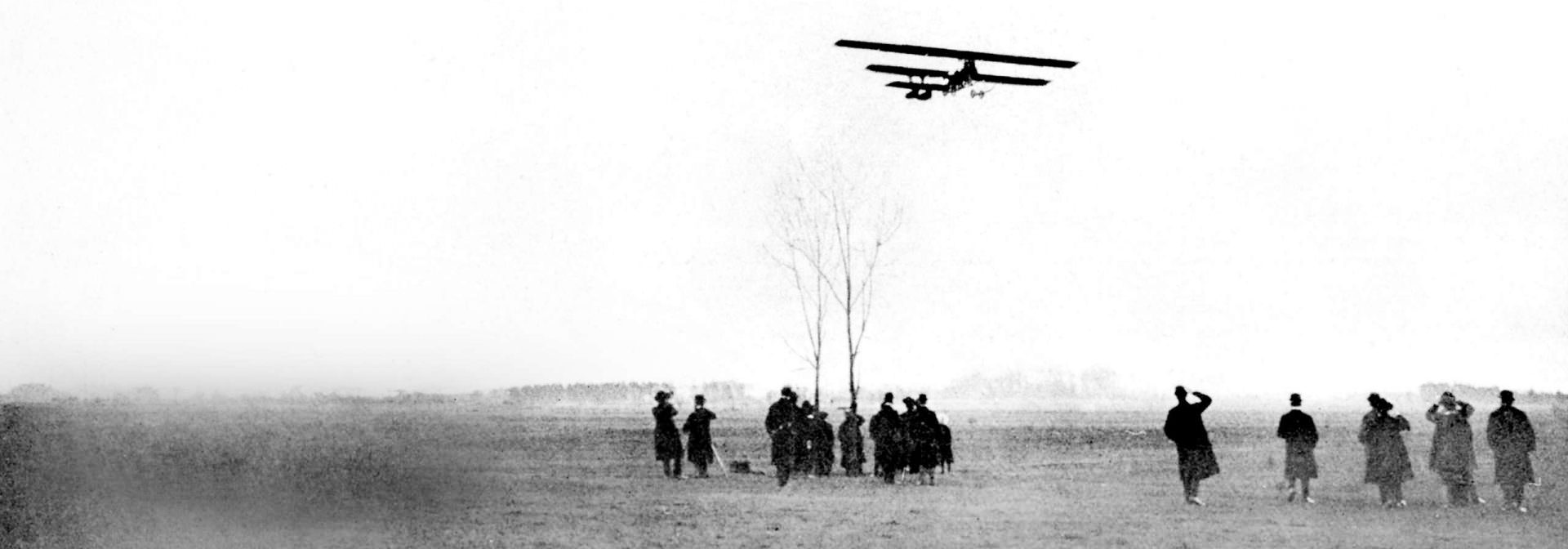 Historic black and white photograph showing a BMW biplane flying low over a field with a group of people watching from the ground.