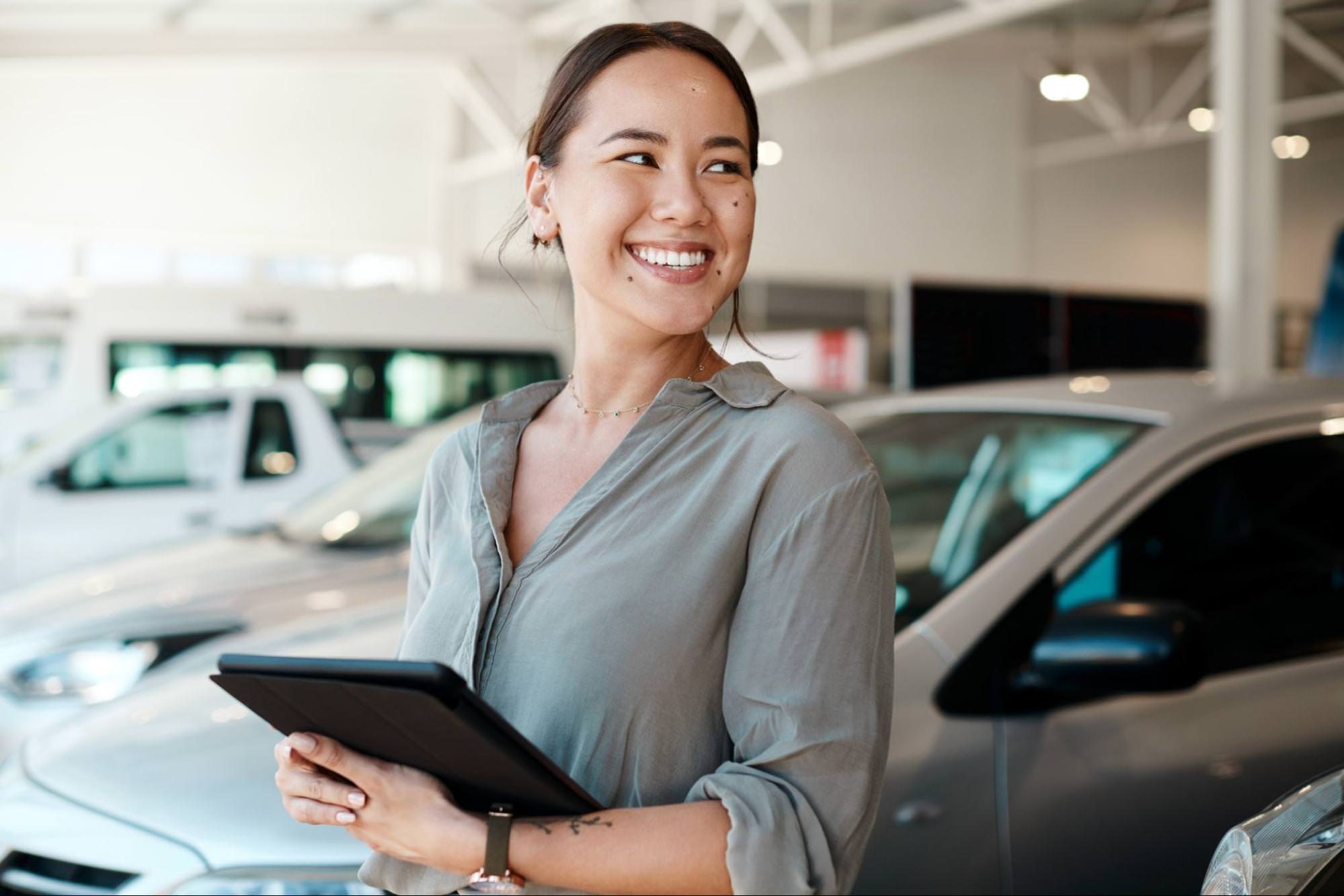 A smiling car salesperson standing in a showroom with cars in the background. She is holding a tablet, appearing friendly and professional, ready to assist customers.