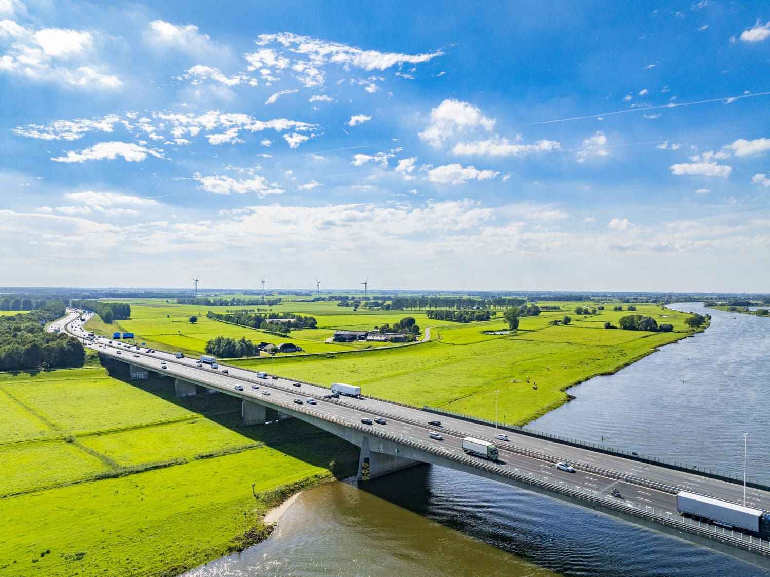 Вид с воздуха на голландский пейзаж с длинным автомобильным мостом через реку. Мост окружен пышными зелеными полями и сельскохозяйственными угодьями. Видно, как по мосту едут машины.