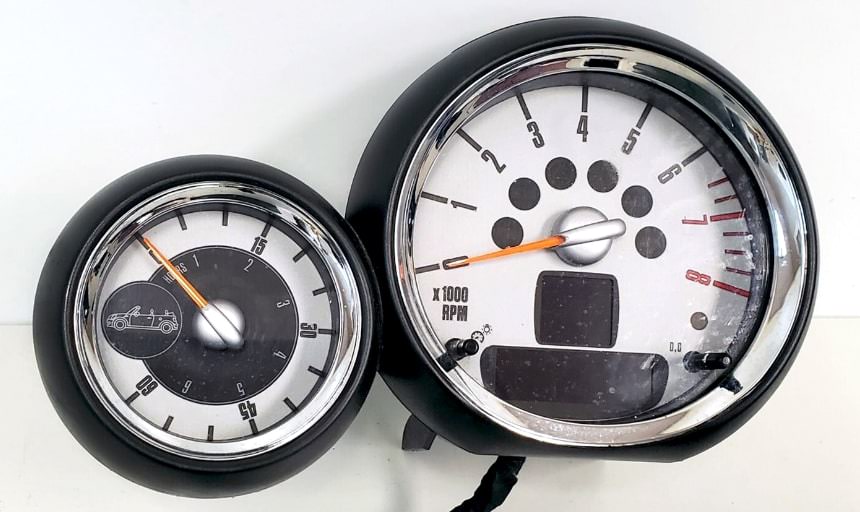 Zwei runde Anzeigen aus dem Armaturenbrett eines Mini Cooper. Die größere Anzeige rechts ist der „Openometer“, eine einzigartige Funktion, die die Fahrzeit mit offenem Verdeck misst. Die kleinere Anzeige links ist ein Standardtachometer. Beide Anzeigen sind in schwarzen Gehäusen untergebracht, die typisch für das retroinspirierte Innendesign von Mini sind.