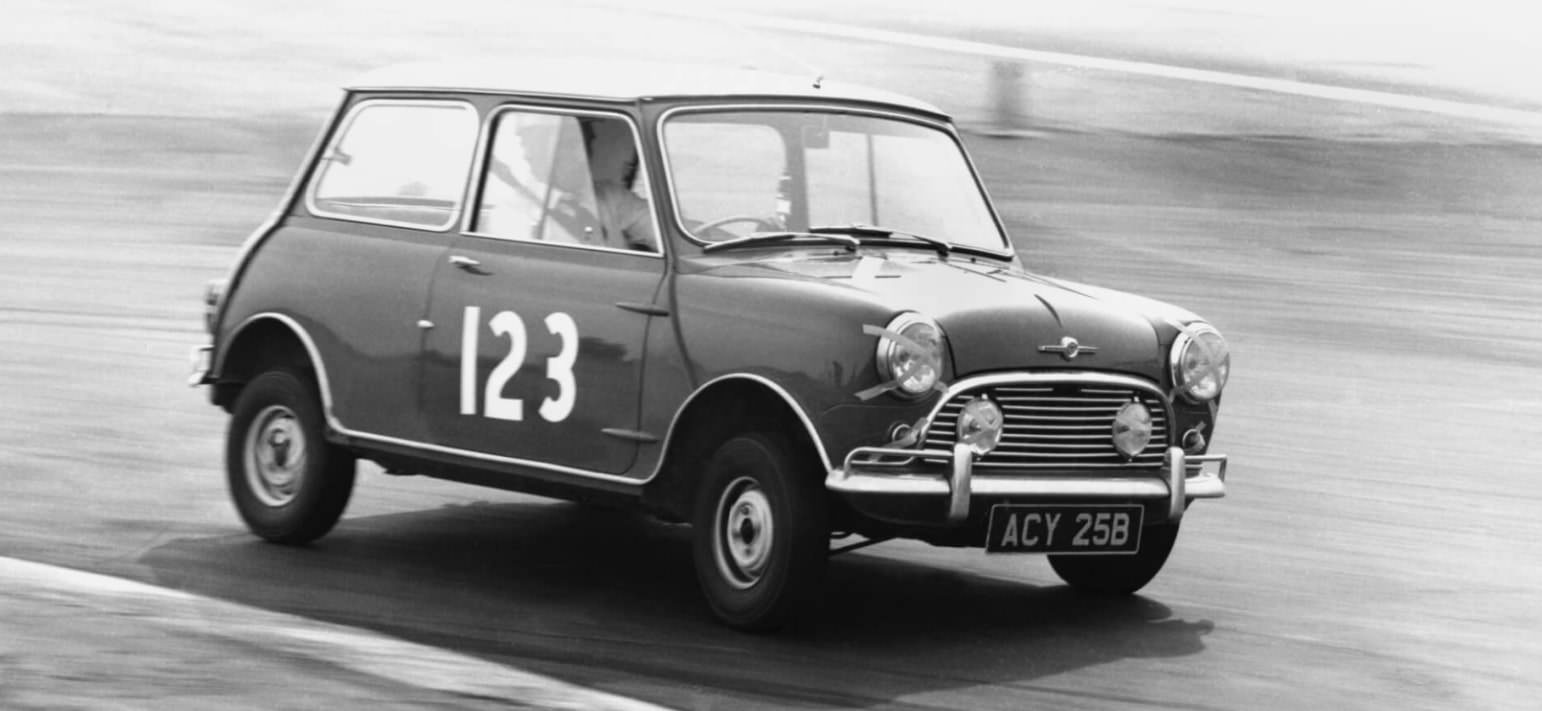 Ein Schwarzweißfoto eines klassischen Mini Cooper-Rennwagens. Das kleine, kastenförmige Fahrzeug ist in Bewegung auf einer Rennstrecke zu sehen, leicht geneigt, als würde es eine Kurve fahren.