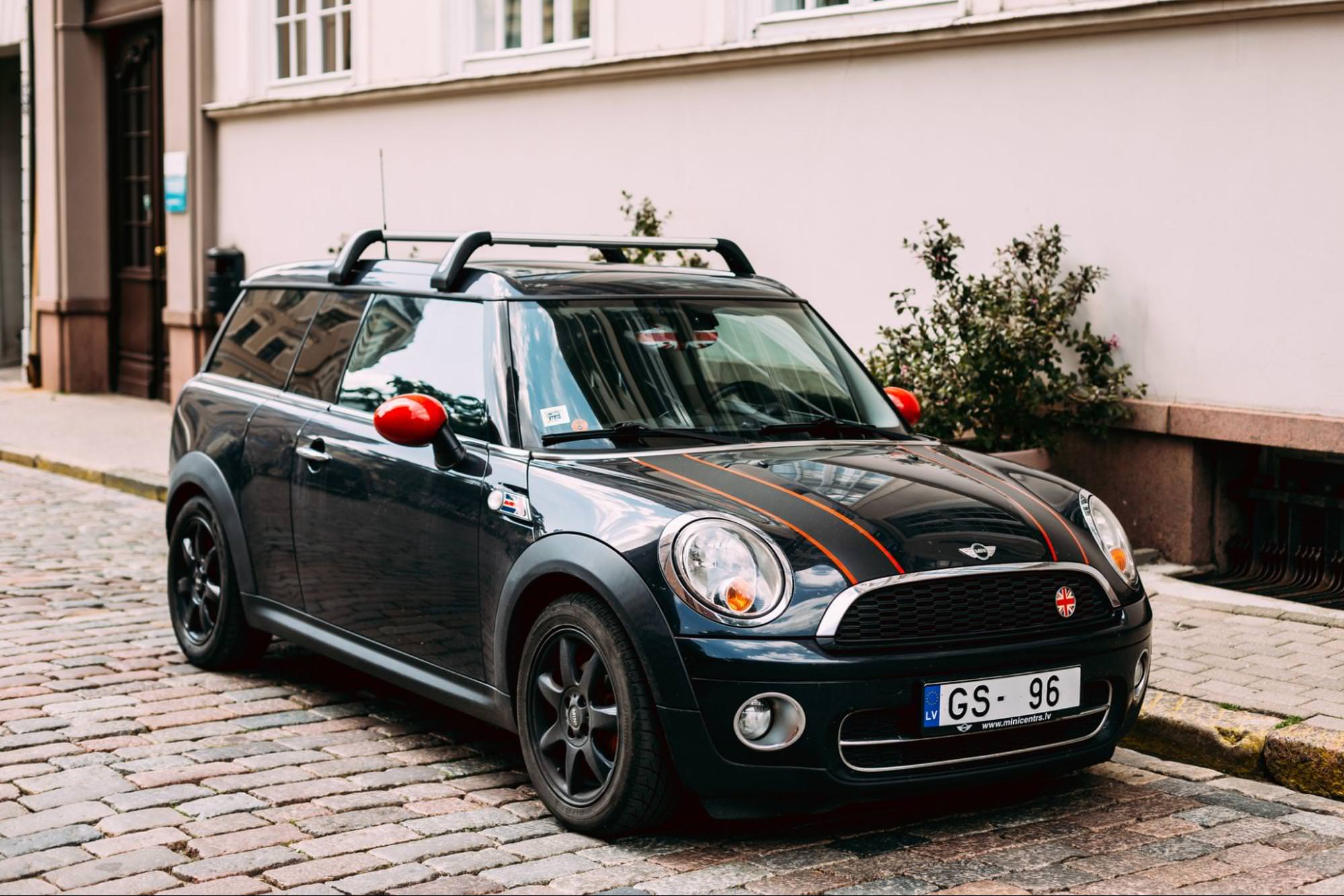 Une Mini Clubman vert foncé garée dans une étroite rue pavée. La carrosserie plus longue et les portes arrière divisées uniques du Clubman sont visibles, ce qui le distingue des autres modèles Mini. Des bâtiments d’aspect historique encadrent l’arrière-plan.