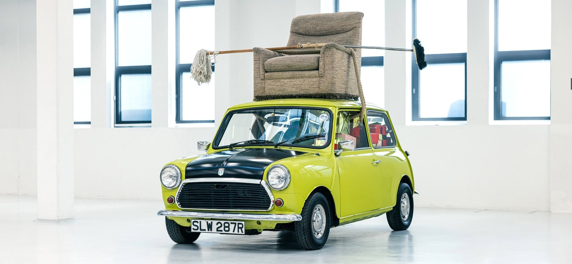 Ein klassischer Mini Cooper in leuchtendem Limettengrün, der an Mr. Beans Kultauto erinnert, steht in einem weißen Innenraum. Das Auto hat ein schwarzes Dach und auf dem Dachgepäckträger ist ein gepolsterter Sessel befestigt, eine Anspielung auf einen berühmten Mr. Bean-Sketch.