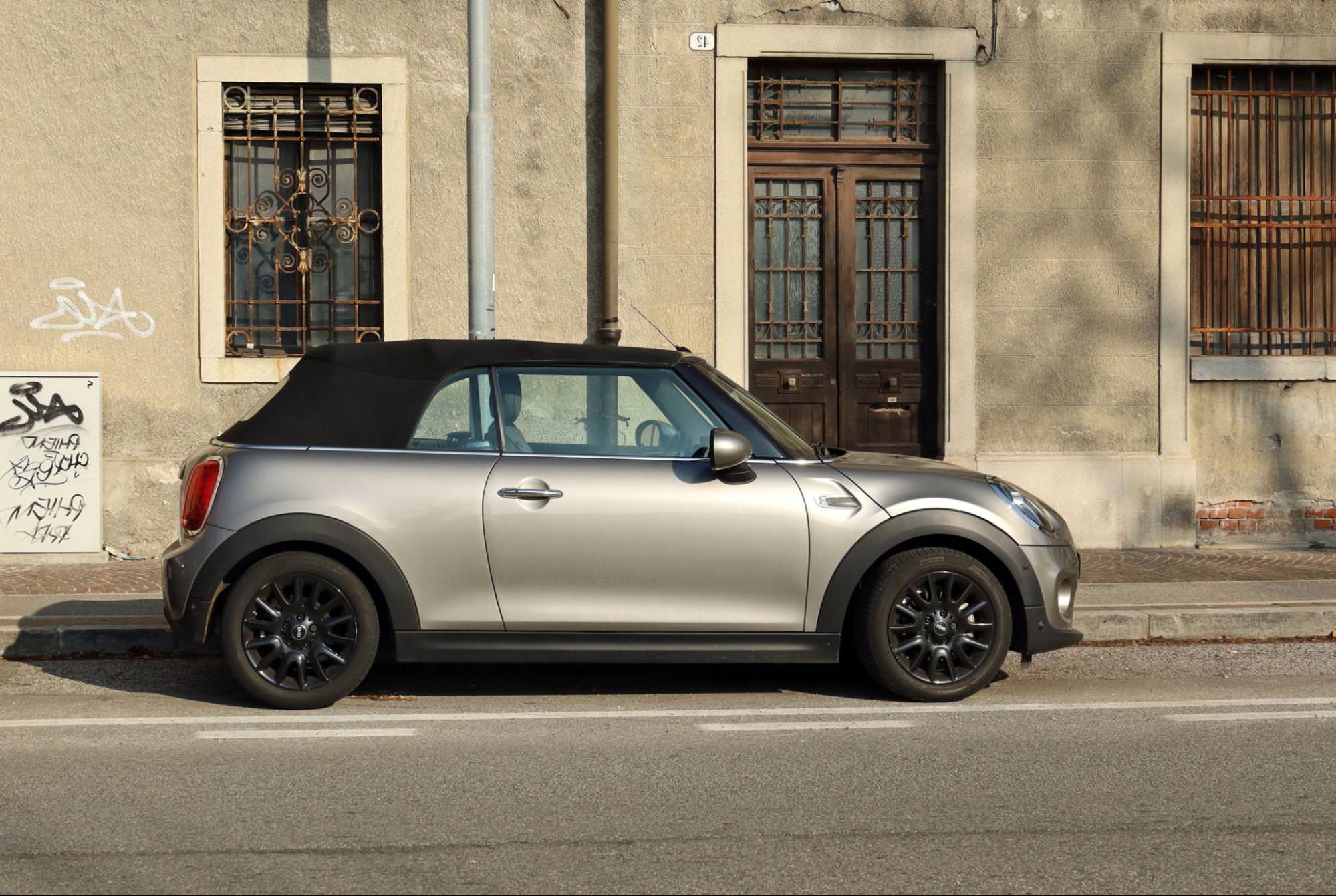 Ein silbernes Mini Cabrio mit geschlossenem Verdeck, geparkt auf einer Stadtstraße. Das Auto steht vor einem hellen Gebäude, was sein schlankes Profil und sein sportliches Design betont.