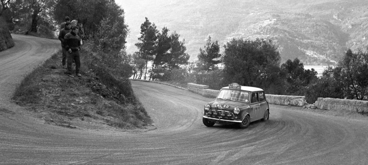 Une photographie en noir et blanc d'un rallye de Monte-Carlo des années 1960. Une Mini Cooper classique est représentée en train de prendre un virage serré sur une route de montagne. Au premier plan, des spectateurs regardent depuis une position élevée au bord de la route.