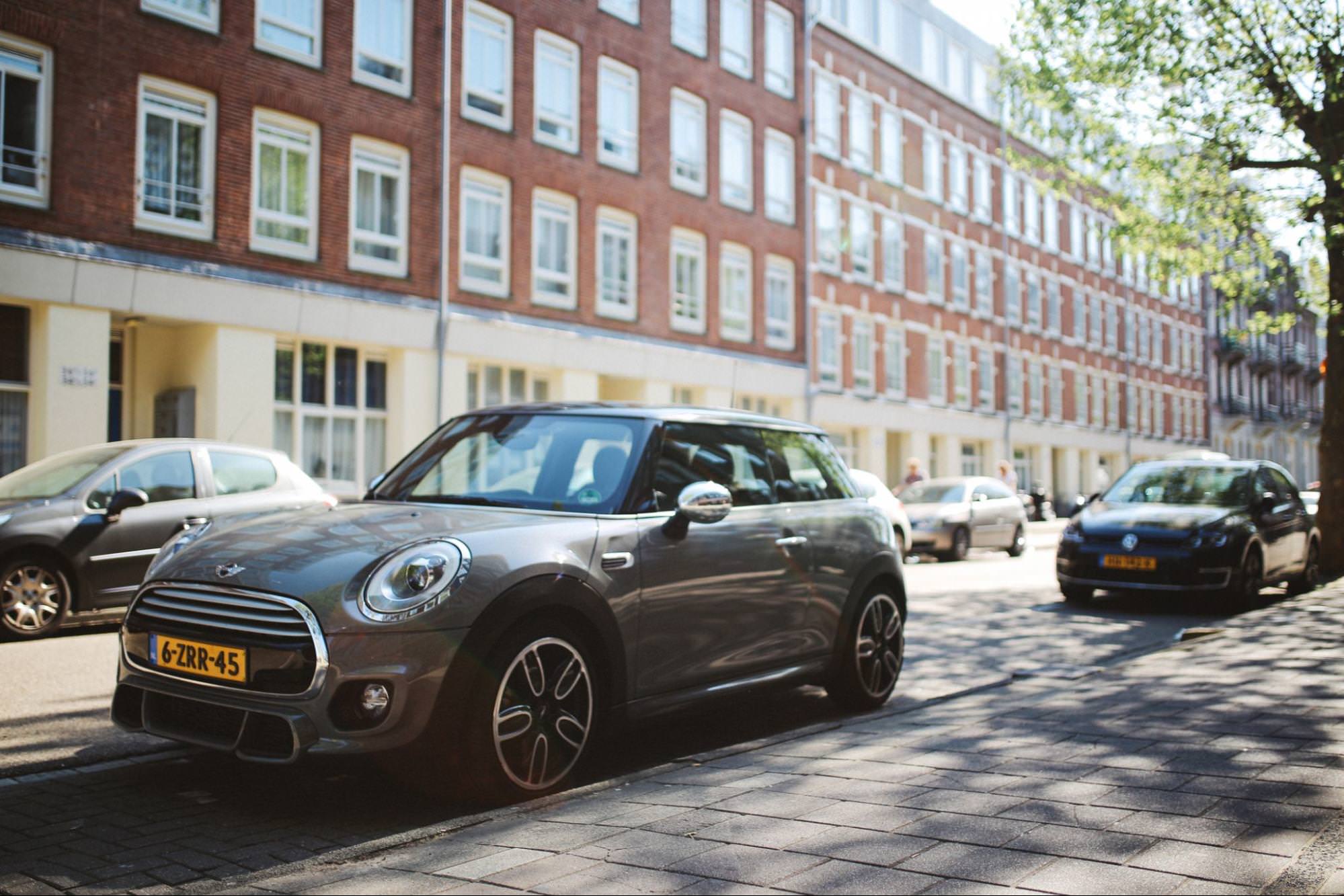 Une Mini Cooper de couleur sombre garée dans une rue bordée d’arbres devant des maisons de ville en brique. D'autres voitures sont visibles en arrière-plan, mettant en valeur la taille compacte de la Mini en milieu urbain.