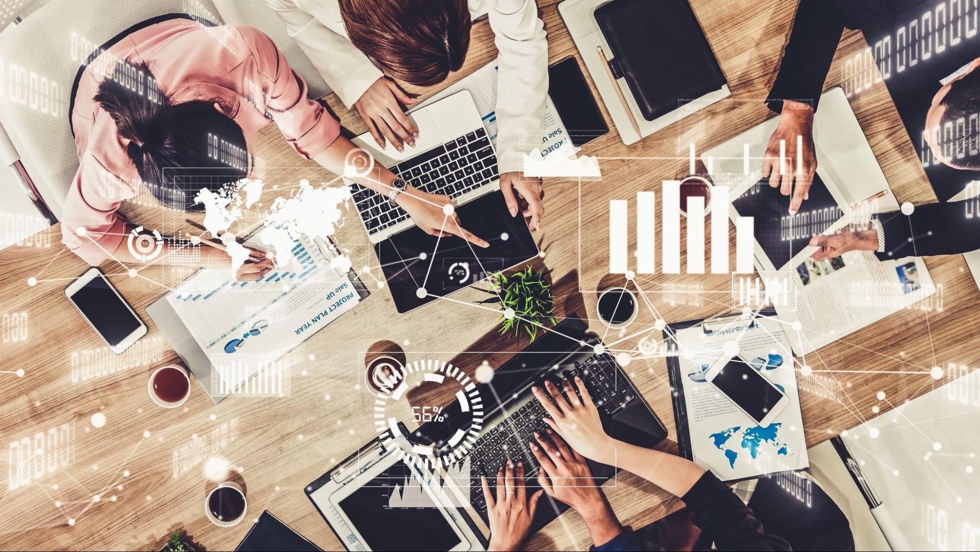 Overhead view of a bustling office desk with multiple hands working on laptops, tablets, and papers. Various charts and graphs are overlaid, suggesting data analysis and teamwork in progress.