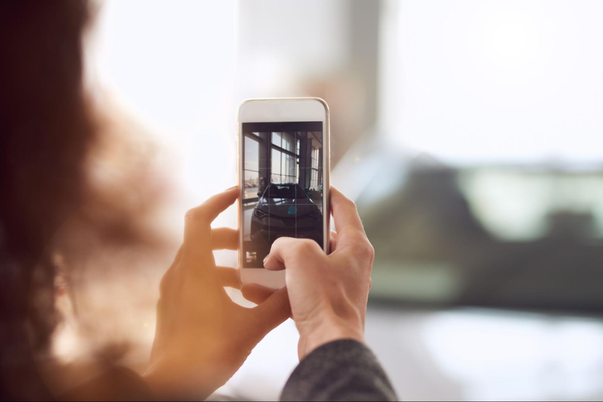 Close-up of hands holding a smartphone, taking a photo of a car inside a showroom. The phone's screen shows the car in focus, demonstrating how to capture engaging images for online listings.