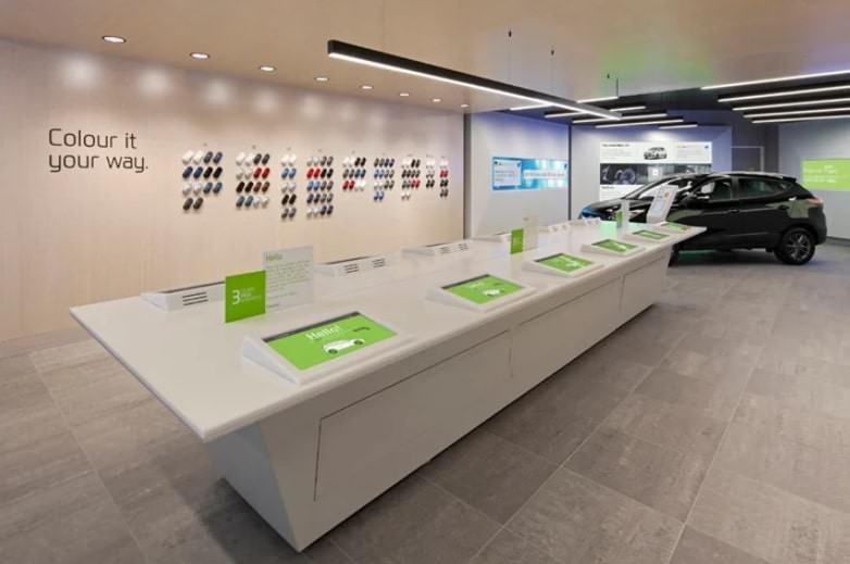 The image shows a modern, interactive car showroom interior. his setup demonstrates an interactive customer experience, allowing potential buyers to explore color options and likely other customization features digitally.