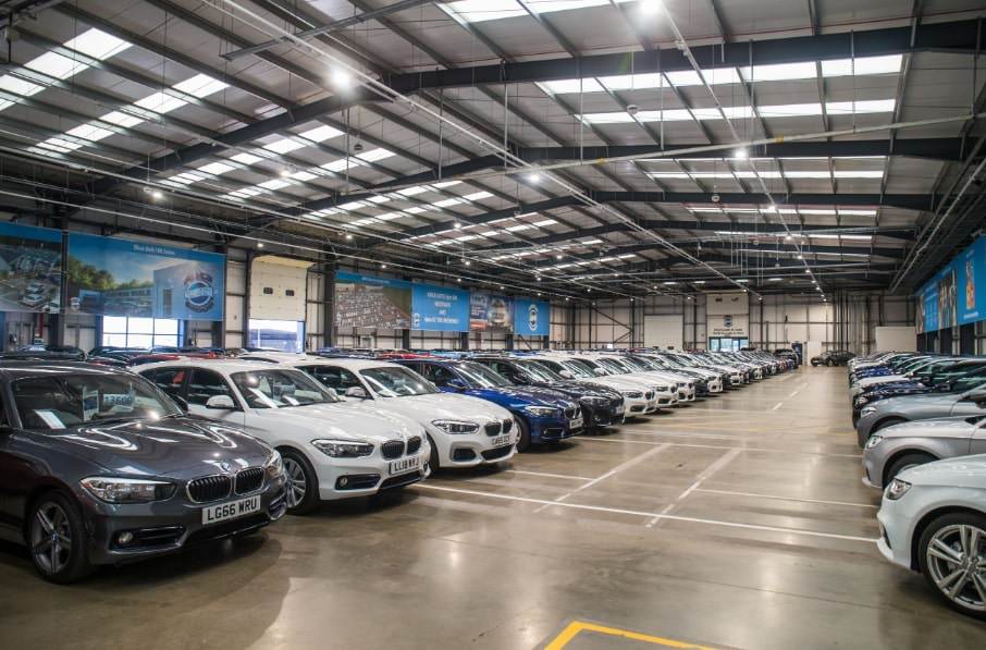 A large, well-lit indoor car showroom with a high ceiling featuring skylights and bright overhead lighting. The spacious floor is filled with rows of clean, shiny cars, predominantly BMWs, neatly arranged in orderly lines.